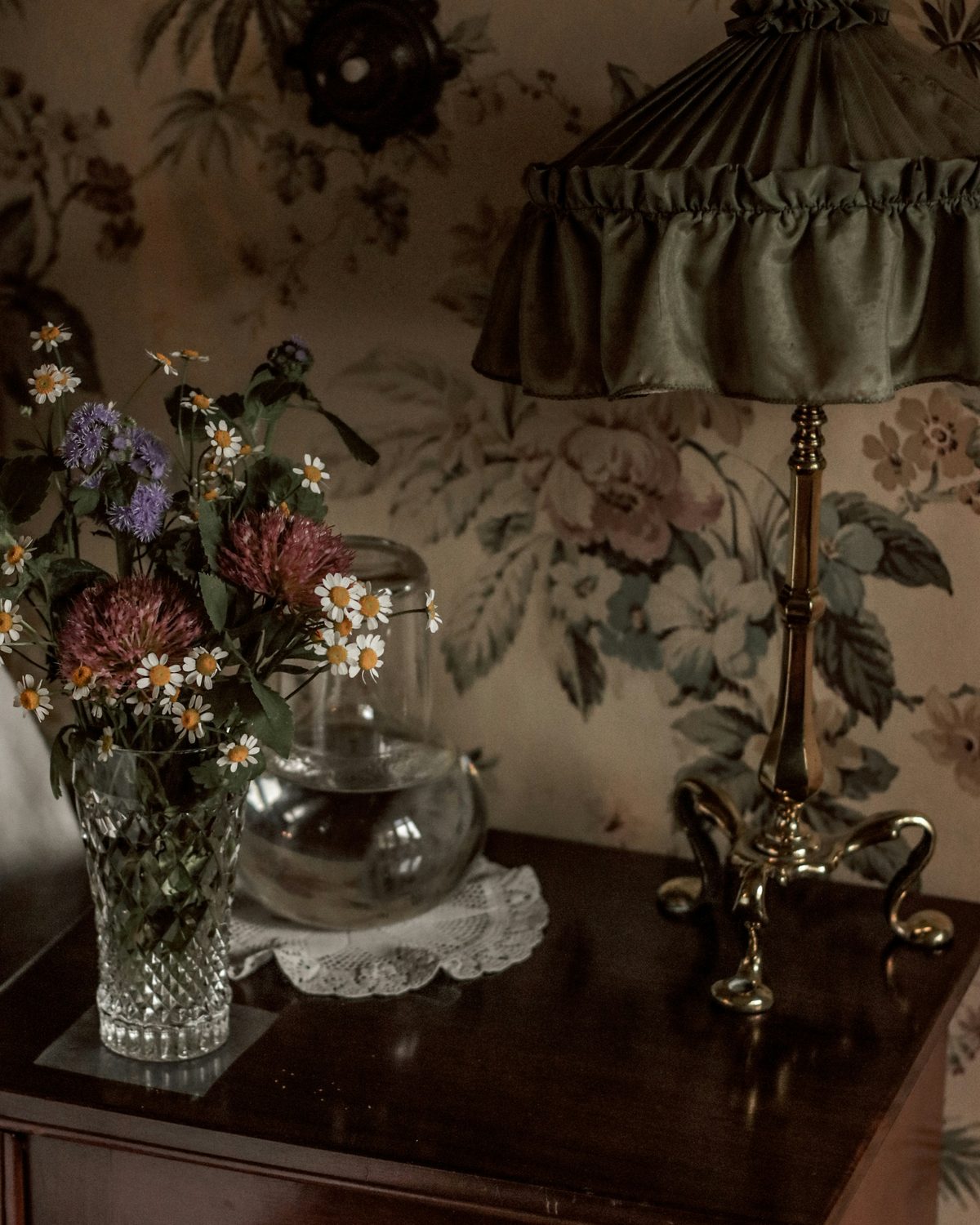 Muted floral wallpaper behind a brass table lamp with a ruffled green shade, a cut-glass vase of wildflowers and a glass carafe on a dark wooden cabinet