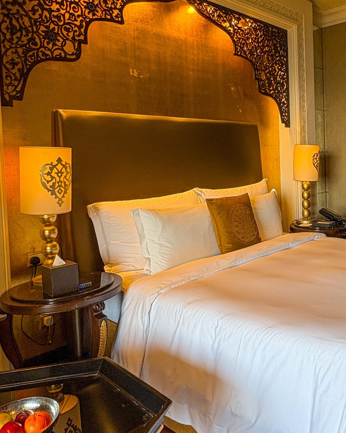 Hotel bedroom with a gold-leaf wall behind the bed, carved fretwork arch, glowing bedside lamps and crisp white linens