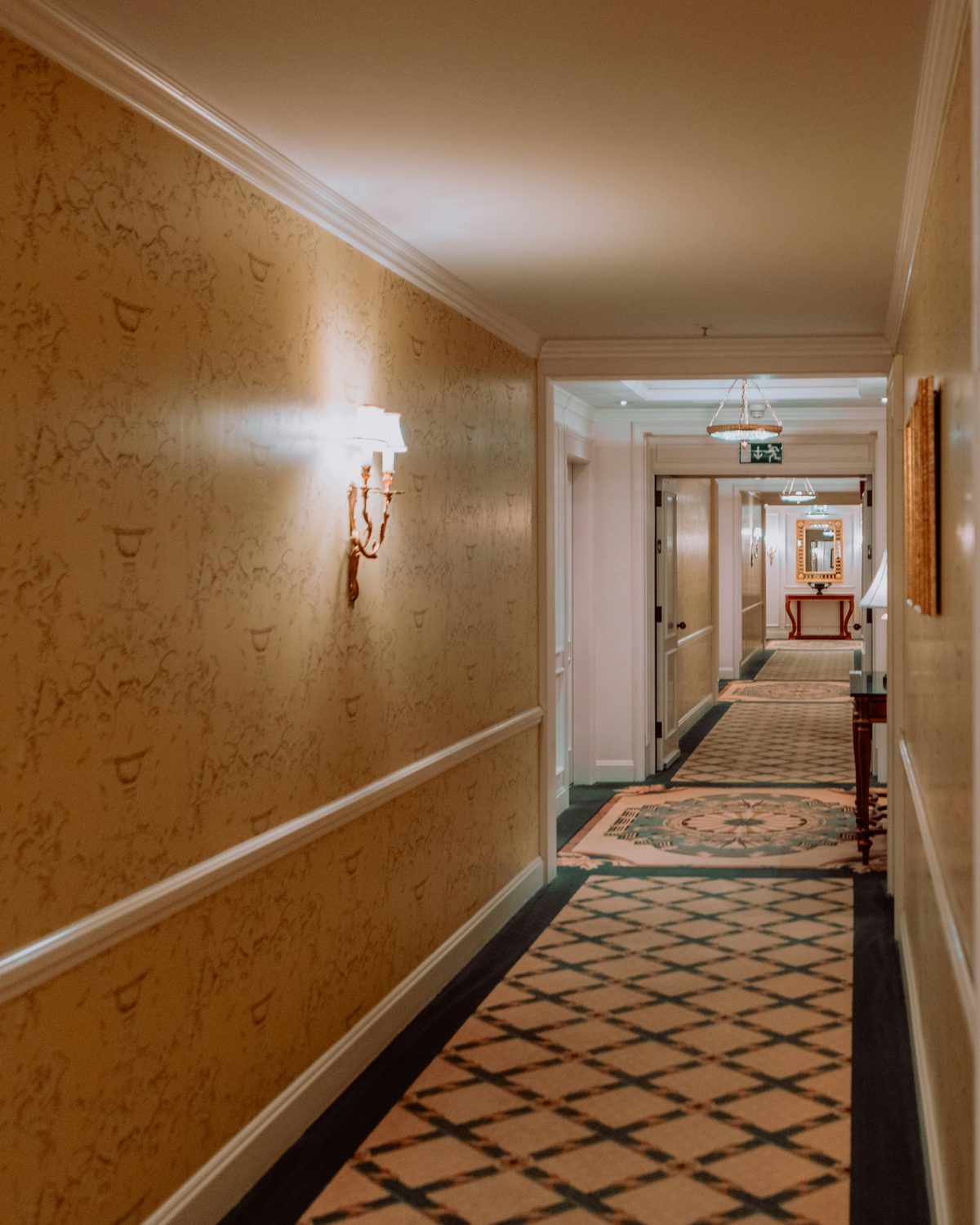 Long hotel corridor with warm gold textured wallcovering, white dado rail, wall sconces and a patterned carpet runner