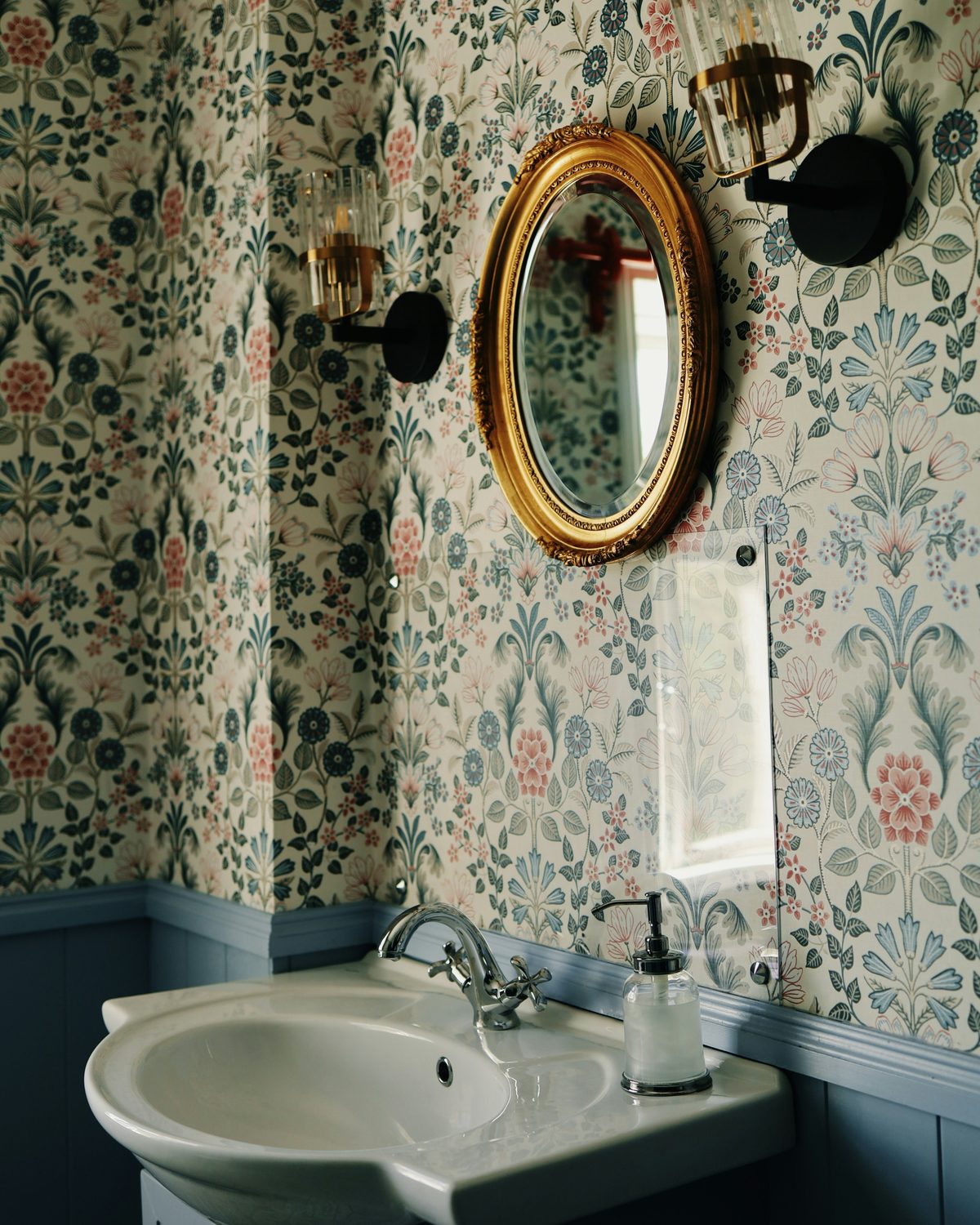 Powder room with a fine floral wallpaper, oval gilt mirror between two wall sconces, and a white basin on a blue vanity