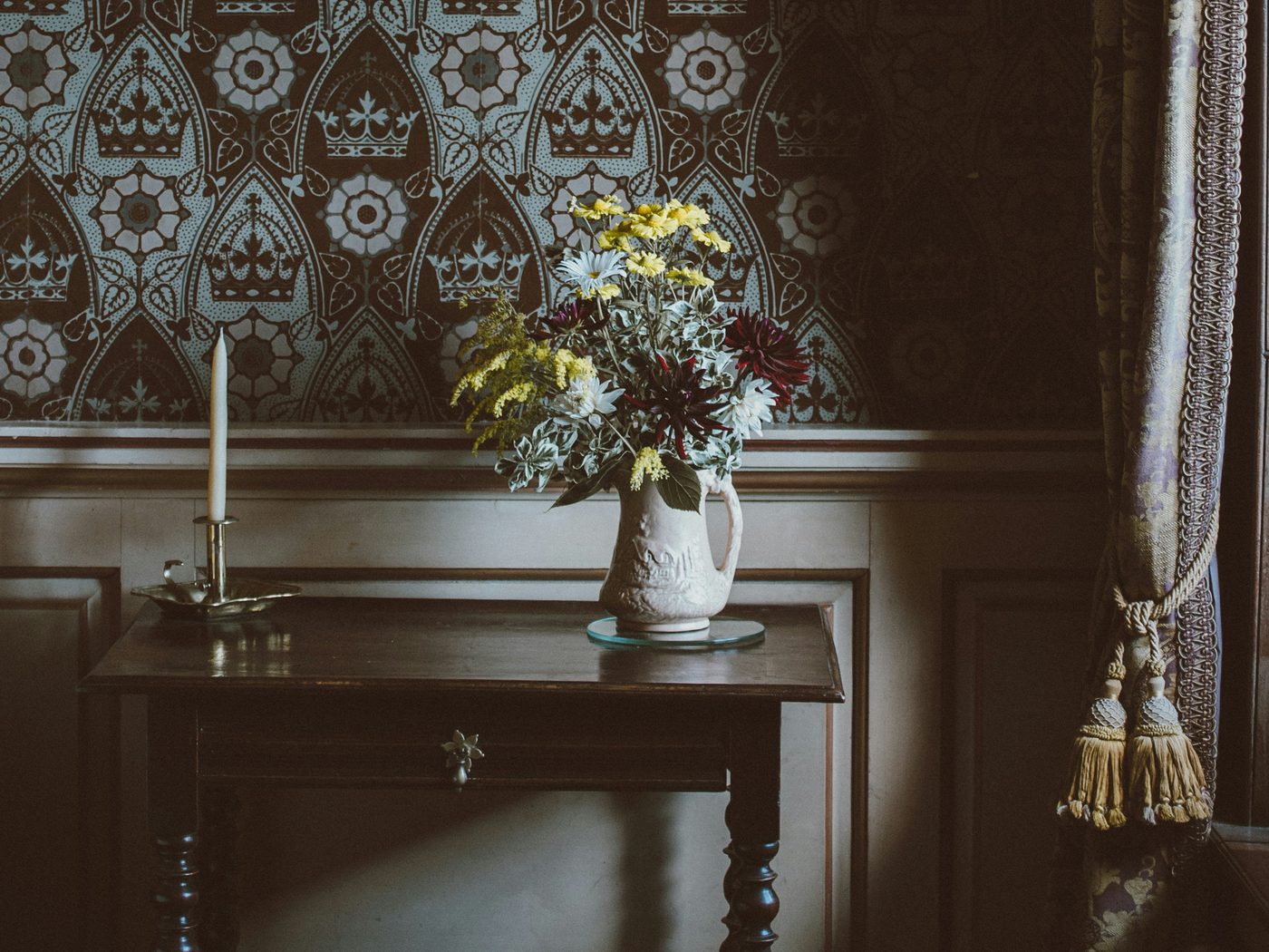 Antique console table with a vase of flowers and a candle against blue-grey crown-and-arch patterned wallpaper above dark panelling
