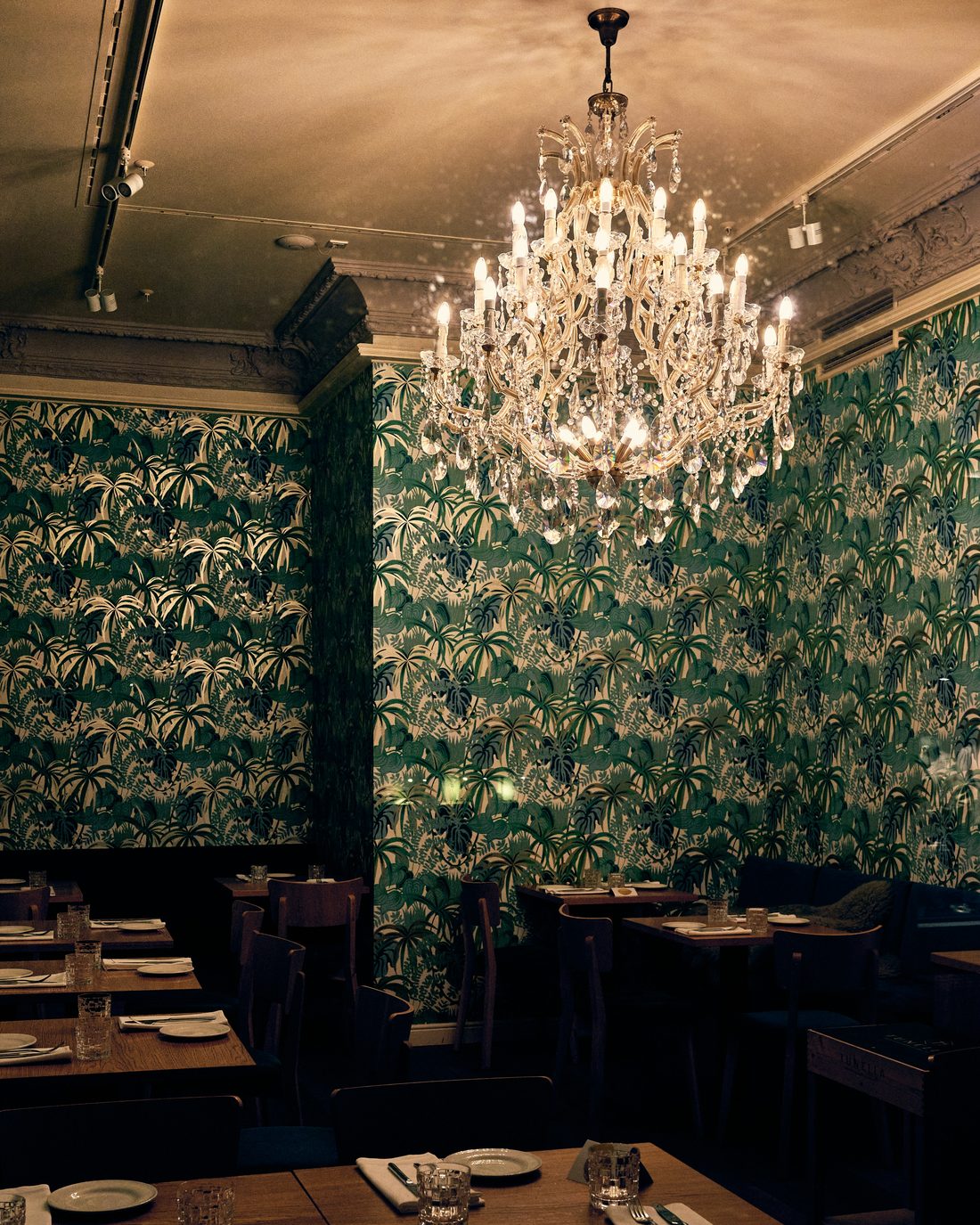 Restaurant dining room wrapped in green palm-leaf wallpaper beneath a large crystal chandelier
